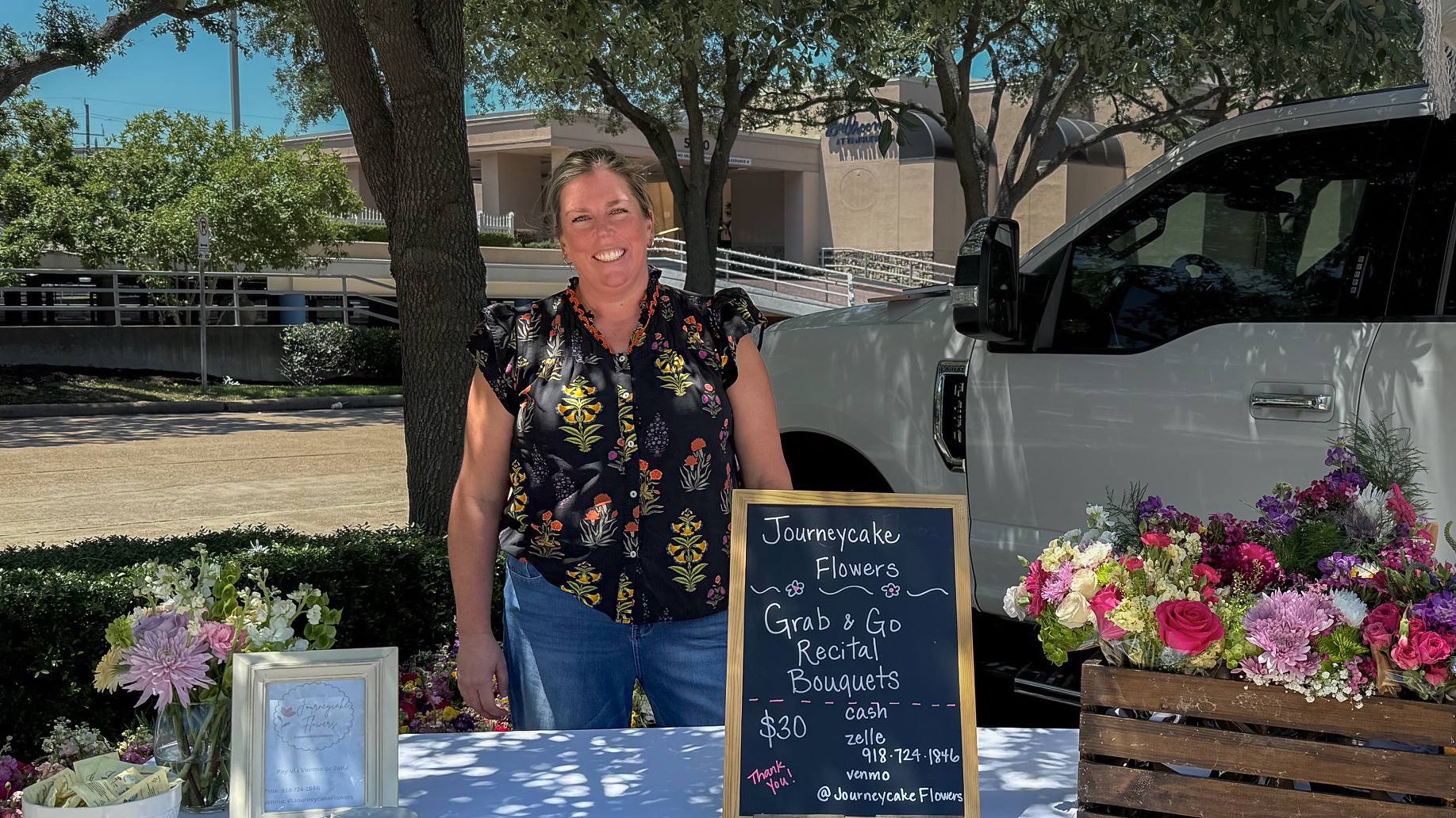 Heather, founder of Journeycake Flowers, smiling behind her flower cart with a Grab & Go bouquets chalkboard sign and a crate of fresh bouquets at an outdoor market