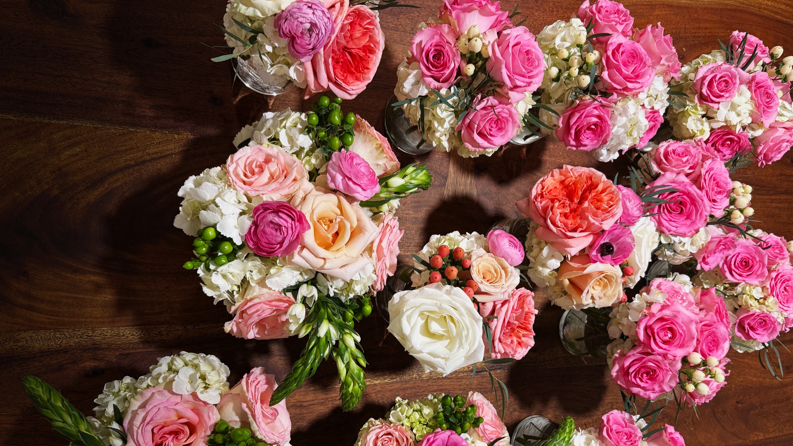An overhead arrangement of fresh-cut bouquets (pink, coral, peach, and white roses, ranunculus, and hydrangea) laid across a warm wood table in soft natural light
