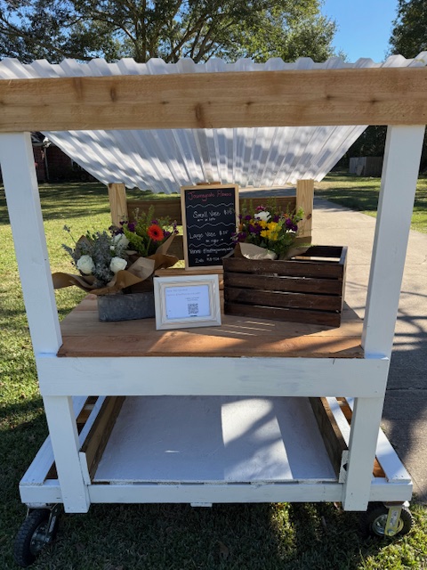 Outdoor white wooden flower cart with a 'Journeycake Flowers' chalkboard sign and two wrapped bouquets